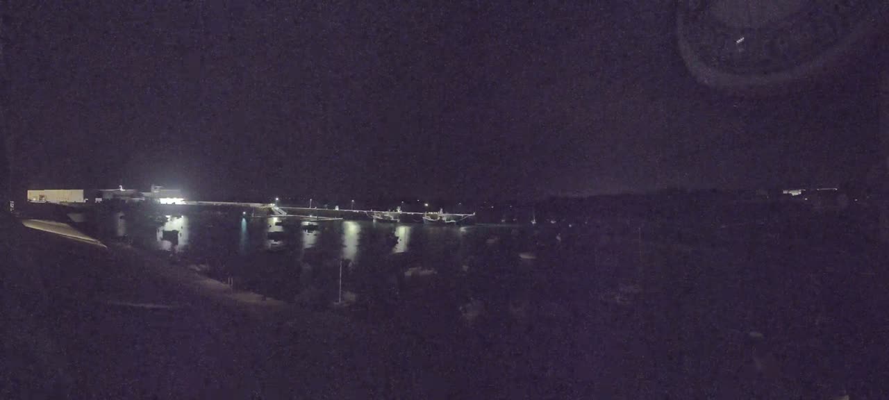 A nighttime view of a harbor with several boats docked, illuminated by dim lights under a dark, clear sky.