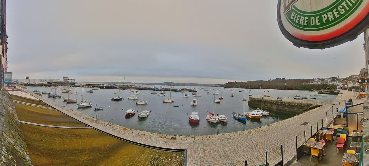 A harbor filled with numerous small boats is seen on an overcast day, with a stone walkway and outdoor seating area in the foreground.