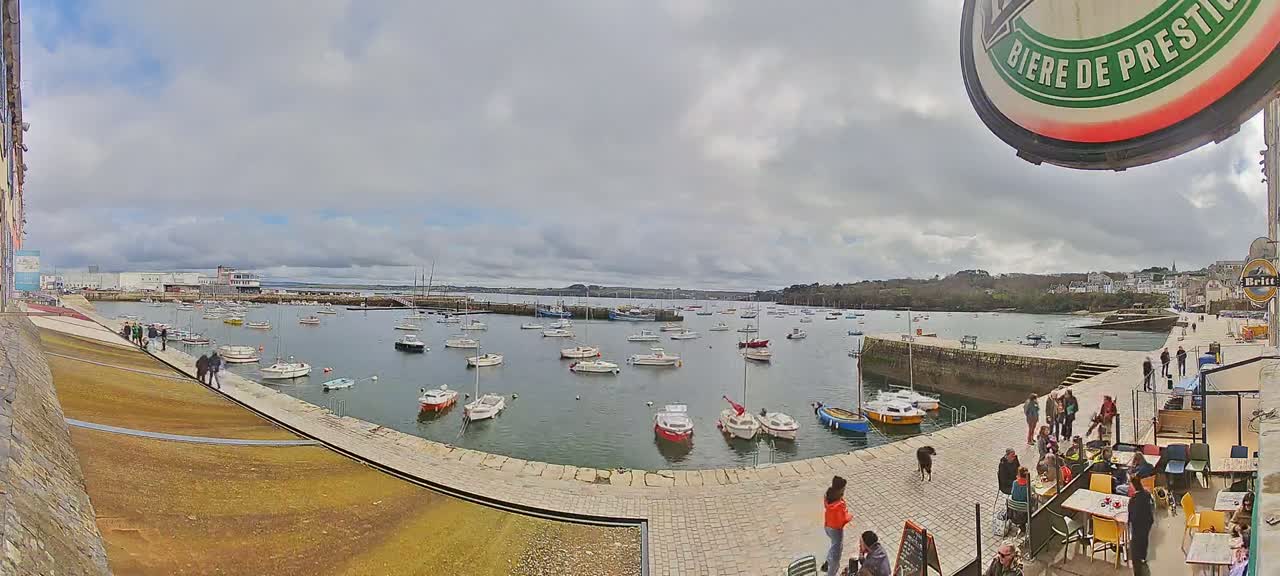 A harbor filled with numerous small boats is viewed from a paved walkway, under a cloudy sky, with people walking along the walkway and sitting at outdoor tables in the foreground.