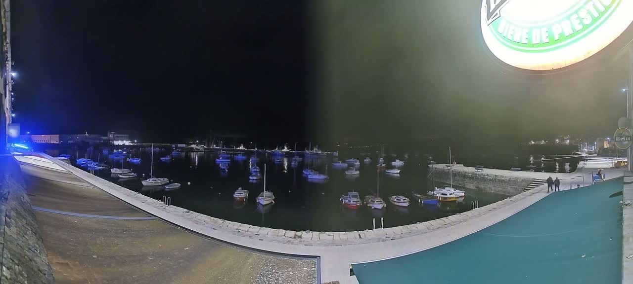 At night, numerous boats are moored in a calm harbor, viewed from a high vantage point overlooking a stone quay.
