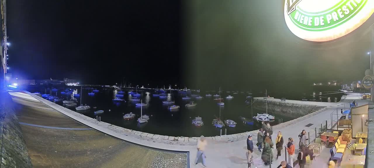 A nighttime view of a harbor filled with small boats, under a dark sky, with several people standing near a stone embankment next to an outdoor seating area.