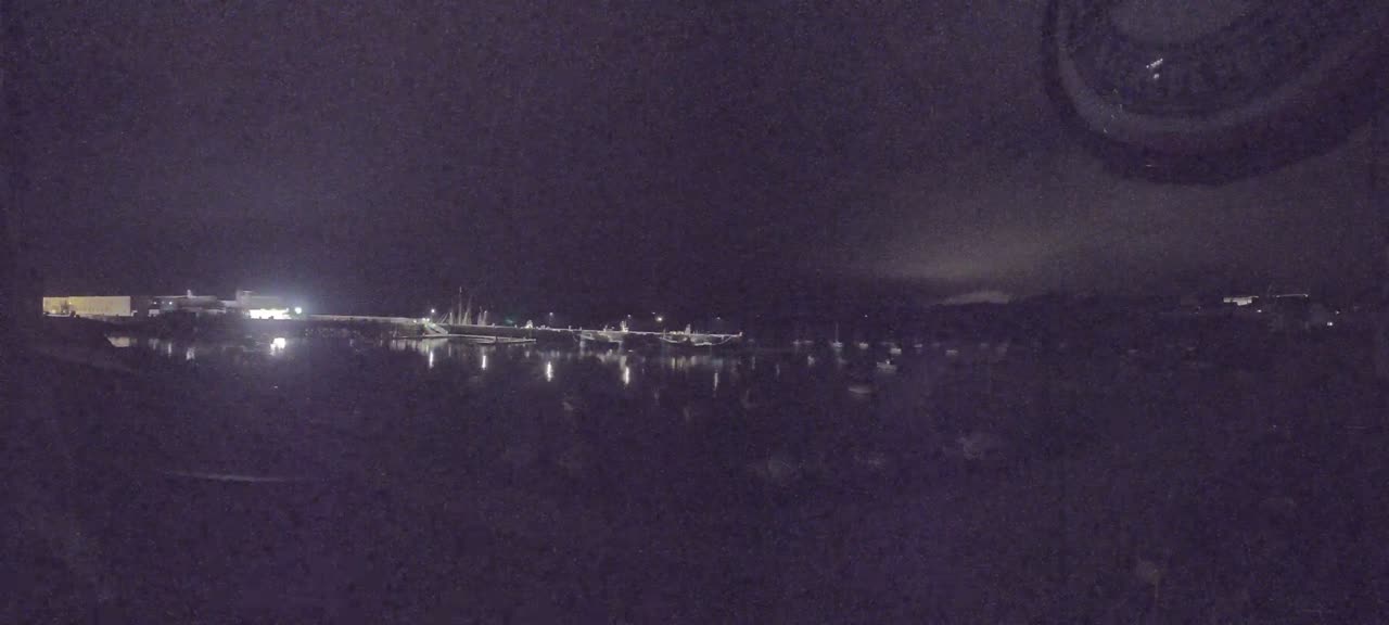 A nighttime view of a calm harbor with several boats moored, illuminated by faint lights, under a dark, cloudy sky.