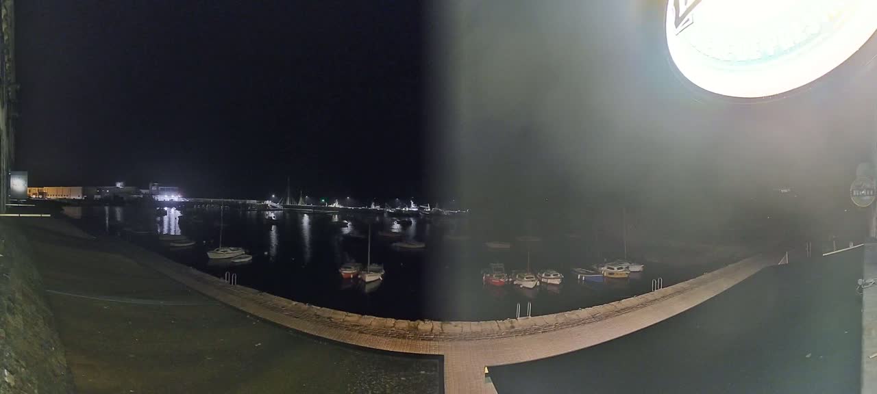 A nighttime view of a harbor filled with small boats, illuminated by lights from buildings and boats on the far side of the water.