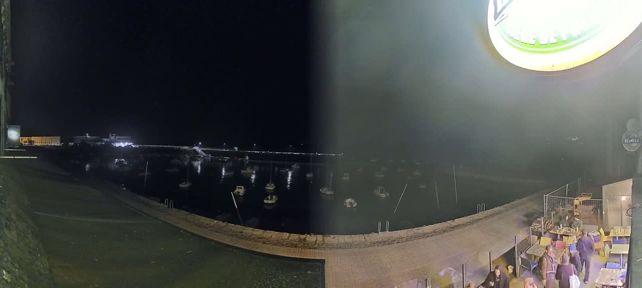 At night, a harbor filled with boats is viewed from a patio area with people seated at outdoor tables.