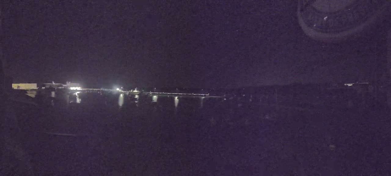 A nighttime view shows a body of water reflecting lights from a distant shoreline under a clear, dark sky.