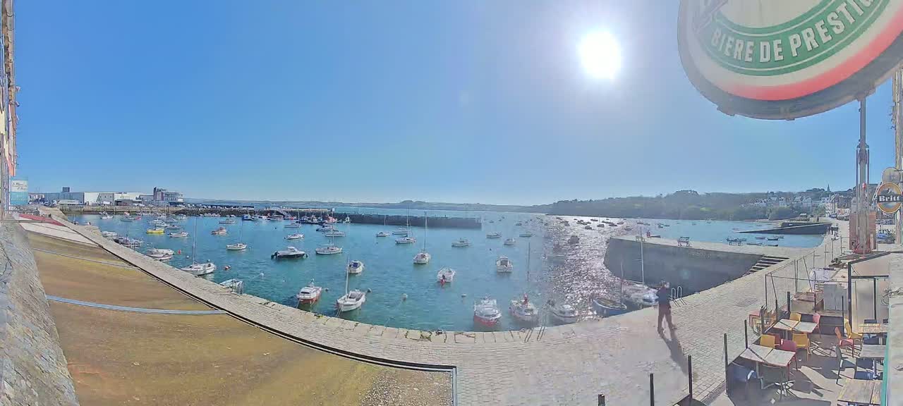 A sunny day shows a harbor filled with numerous sailboats and motorboats, docked along a stone quay with a paved walkway and outdoor seating area.