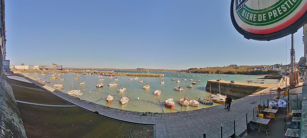 A sunny day reveals a calm harbor filled with numerous small boats, a stone walkway leading to an outdoor cafe, and a distant shoreline.