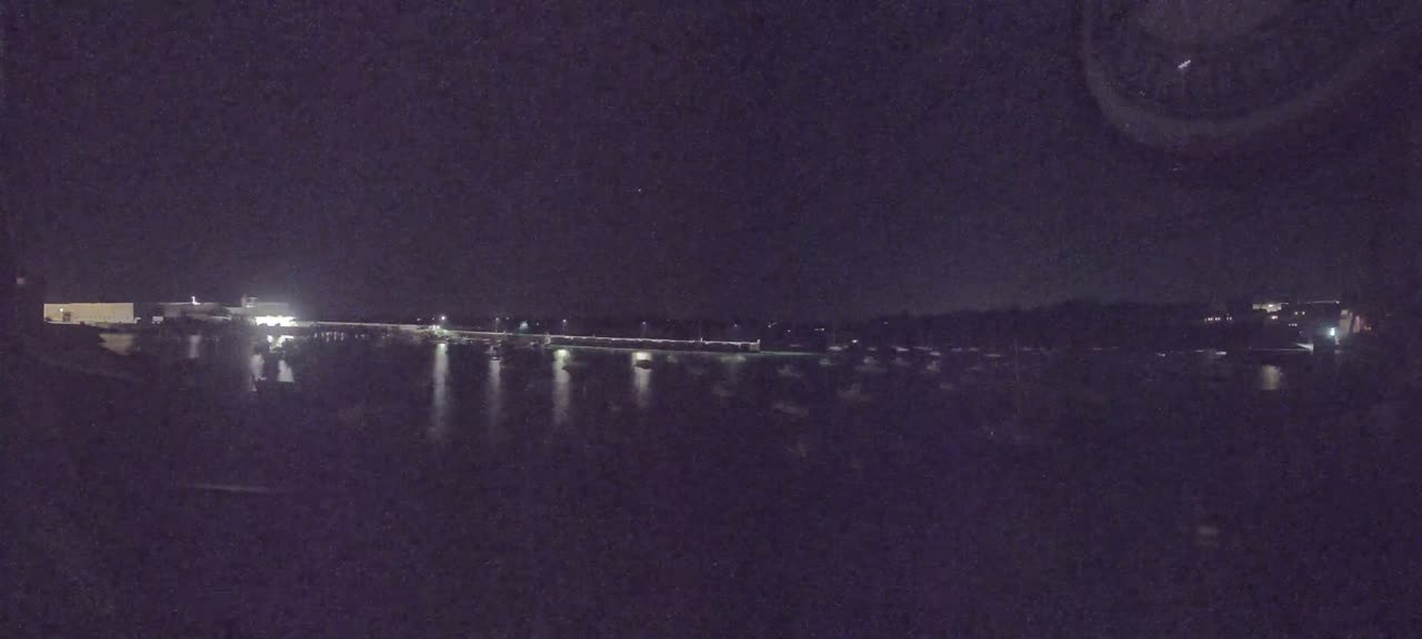 A nighttime view of a calm body of water reflecting the lights from buildings and boats along the shore under a dark, clear sky.