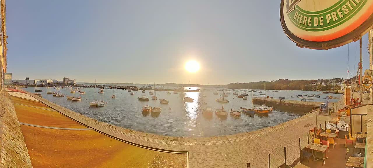 A sunny harbor scene shows numerous boats at rest in calm water, with a stone walkway and outdoor seating area in the foreground.