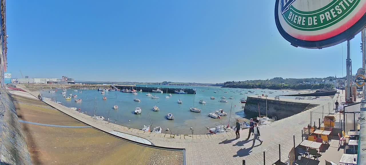 A sunny harbor scene shows numerous small boats moored in calm water, with a few people walking along a paved waterfront area.