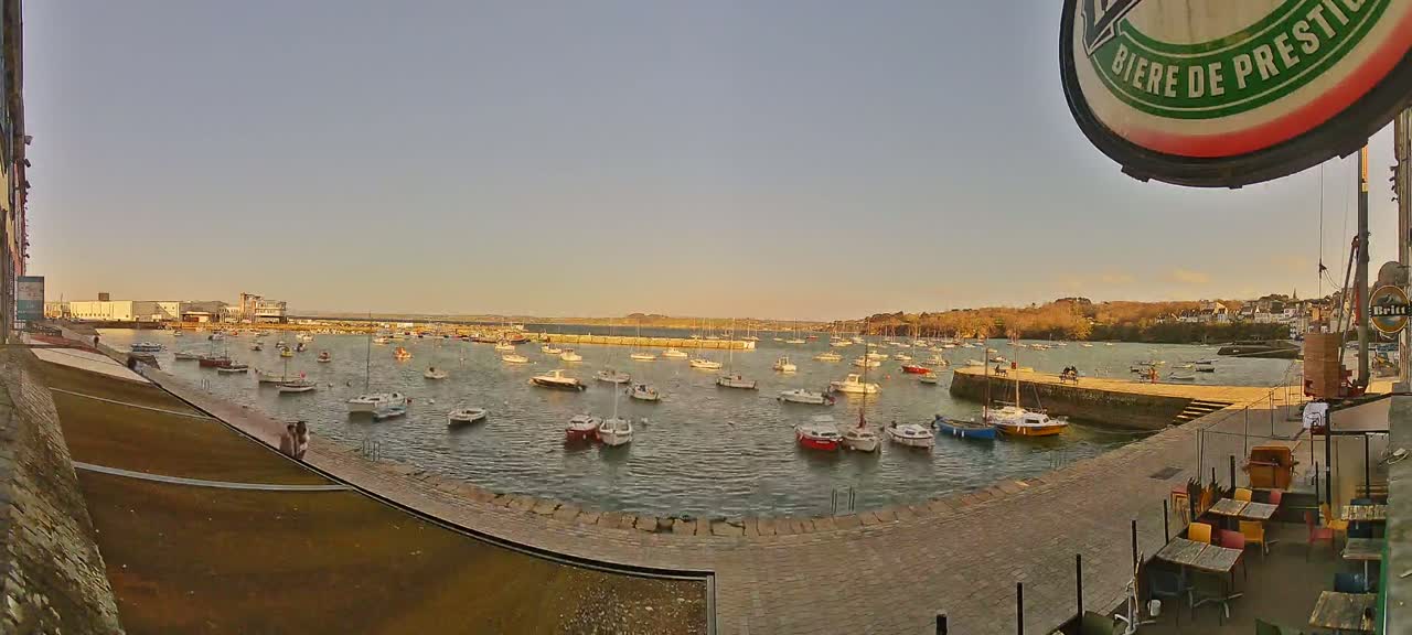 Many small boats are moored in a calm harbor under a clear, pale-yellow sky, with a waterfront patio visible in the foreground.
