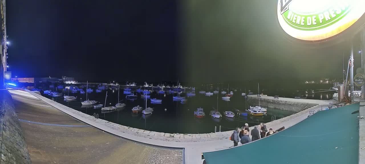 Many small boats are moored in a harbor at night, with a group of people standing on a nearby wall.