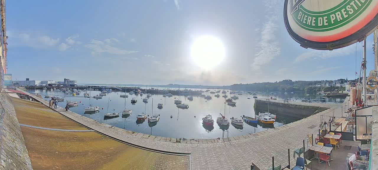A sunny day overlooking a harbor filled with numerous small boats, with a paved area and outdoor seating in the foreground.