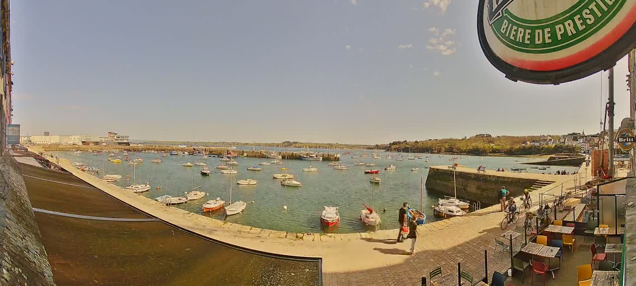 A sunny day reveals a harbor filled with numerous small boats, docked along a stone quay where several people stroll near outdoor seating at a cafe.