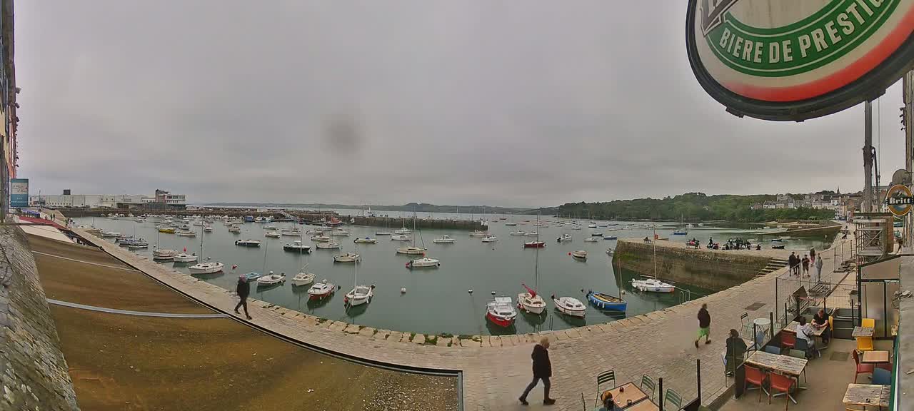 Numerous small boats are moored in a calm harbor on an overcast day, with people strolling along a stone quayside next to outdoor seating at a cafe.