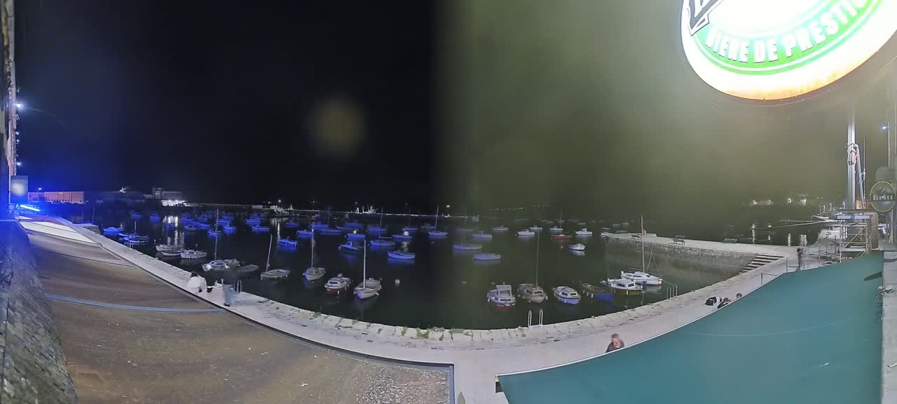A nighttime view of a harbor filled with numerous boats, calm water reflecting the lights, and a few people walking along a nearby stone walkway.