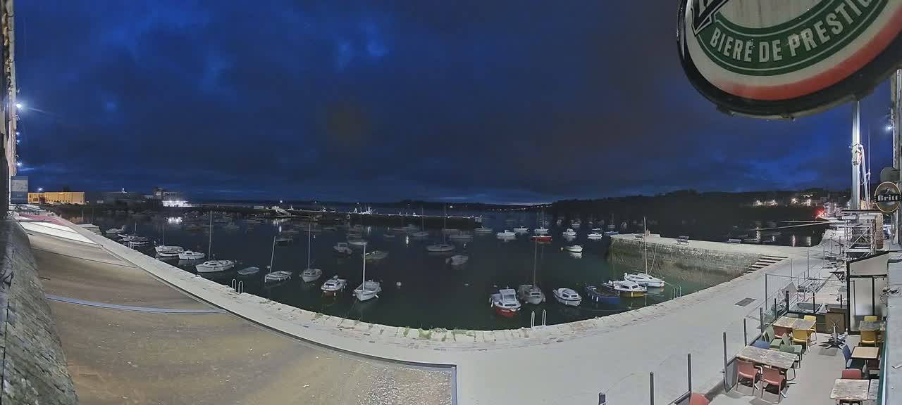 Under a dark blue twilight sky, a harbor filled with numerous small boats is viewed from an elevated position overlooking a paved waterfront area with outdoor seating.