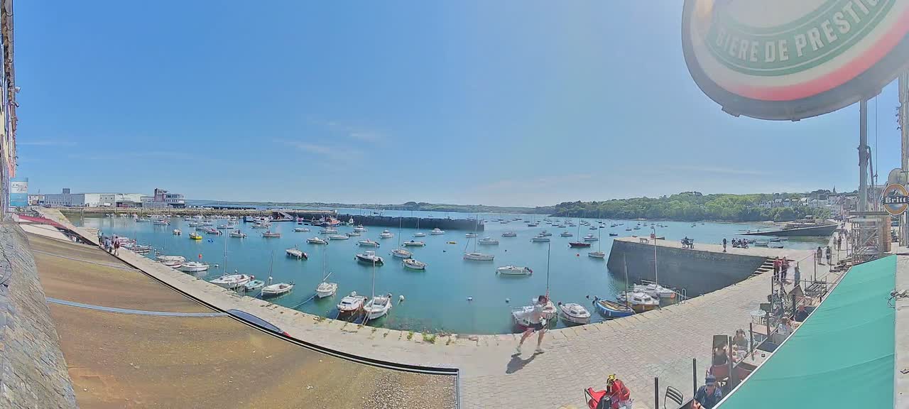 A sunny day at a harbor shows numerous small boats at rest in calm water, surrounded by stone walls and a few people strolling along the waterfront.