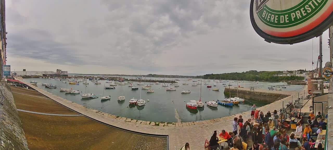 A harbor filled with numerous sailboats is seen under a cloudy sky, with people gathered on a nearby paved area.