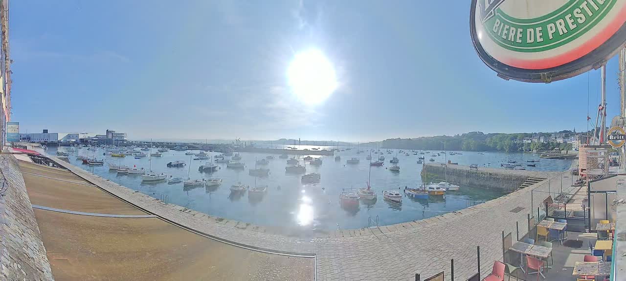 A sunny day shows a harbor filled with numerous small boats, a stone quay, and buildings in the background.
