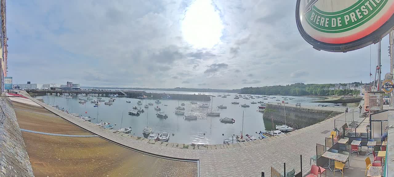 A partly sunny day overlooking a harbor filled with numerous boats, a stone quay, and buildings in the distance.