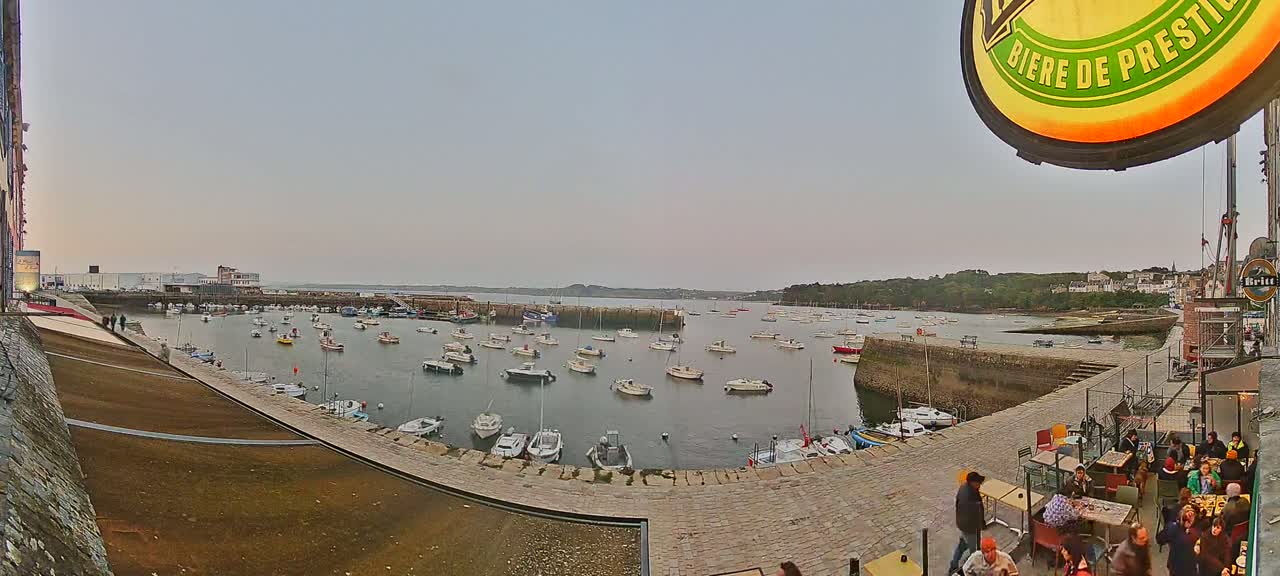 A harbor filled with numerous boats is viewed from an elevated vantage point on an overcast day, with people seated at outdoor tables in the foreground.