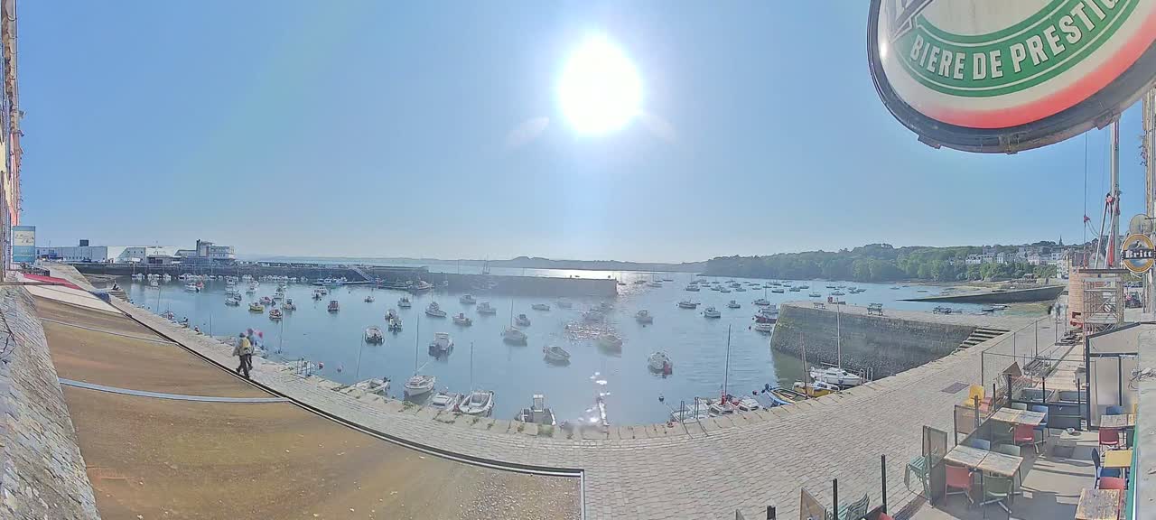 A sunny day overlooks a harbor filled with numerous boats, a stone quay, and buildings in the background.