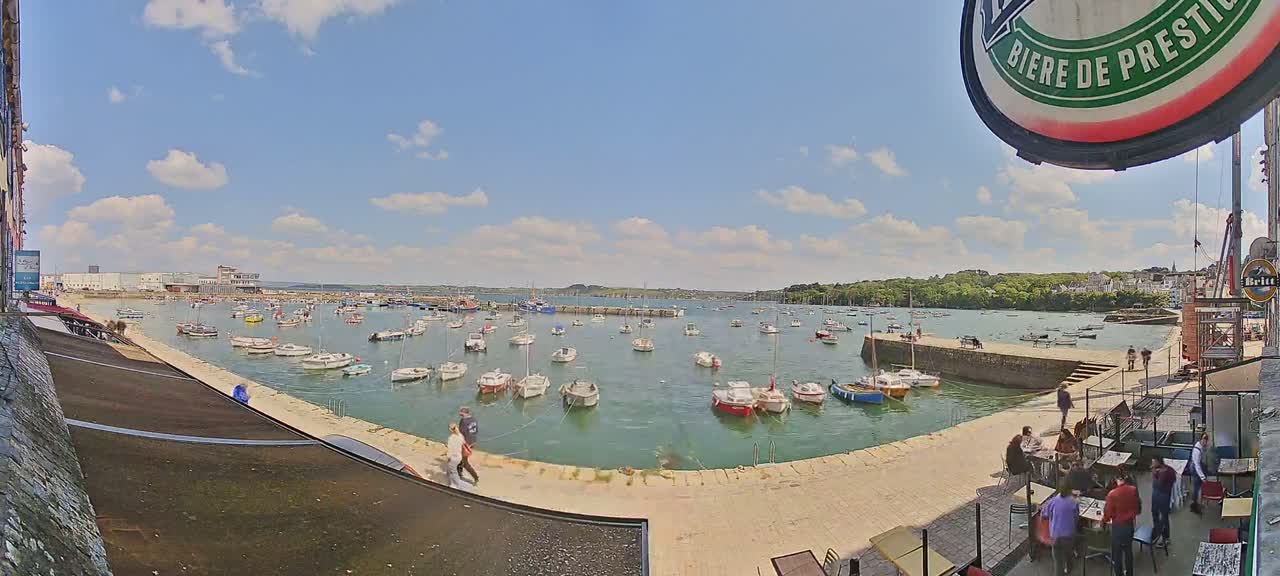 A harbor filled with numerous small boats under a partly sunny sky, with people walking along a paved area near an outdoor restaurant.