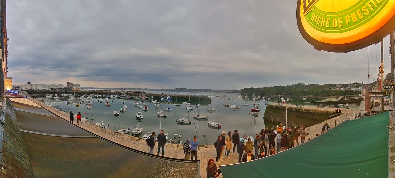 A harbor filled with numerous boats is viewed from a waterfront location under an overcast sky, with a crowd of people gathered on a stone walkway.