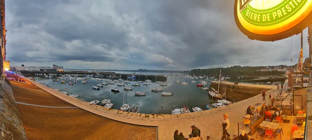A harbor filled with numerous sailboats and motorboats is viewed from an elevated position on an overcast day, with a paved walkway and outdoor seating area visible in the foreground.