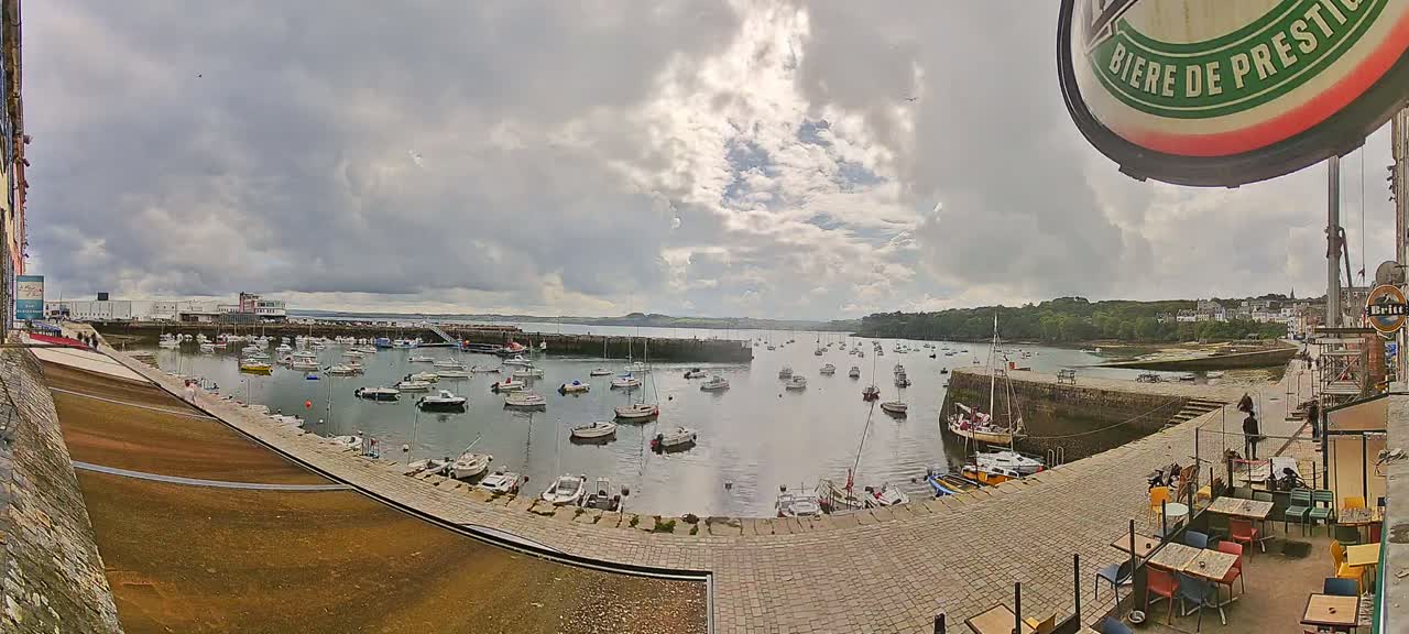 A harbor filled with numerous small boats is seen under a partly cloudy sky, with a paved waterfront area and outdoor seating visible in the foreground.