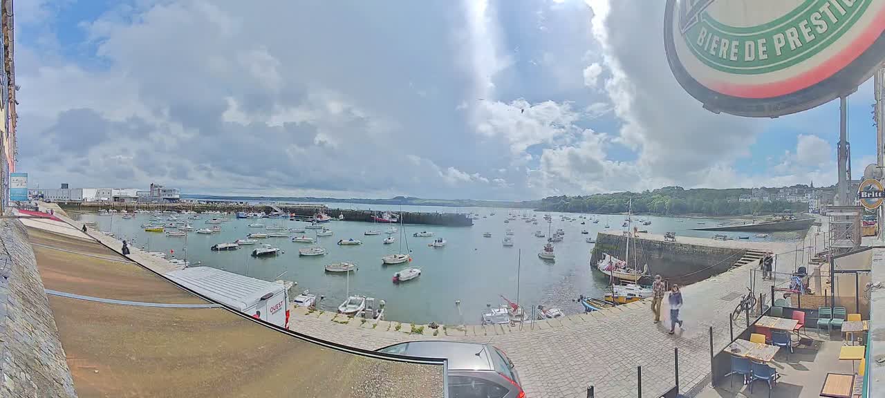 A partly cloudy day reveals a harbor filled with numerous sailboats and motorboats, overlooked by a stone quayside with outdoor seating.