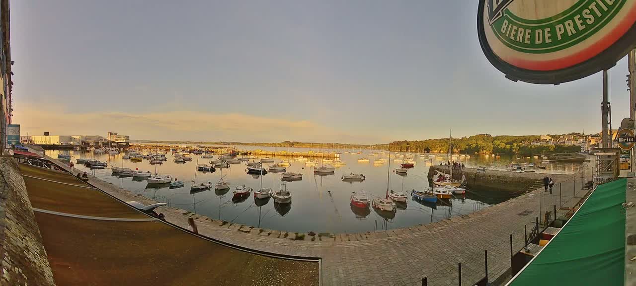 A calm harbor filled with numerous sailboats is viewed from a seaside location under a clear, pale-yellow sunset sky.