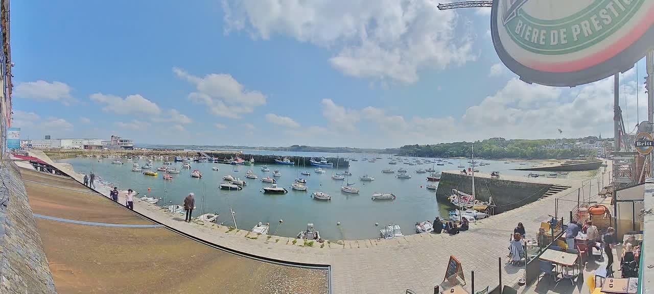 A harbor filled with numerous boats under a partly cloudy sunny sky, with people gathered on a nearby quay.