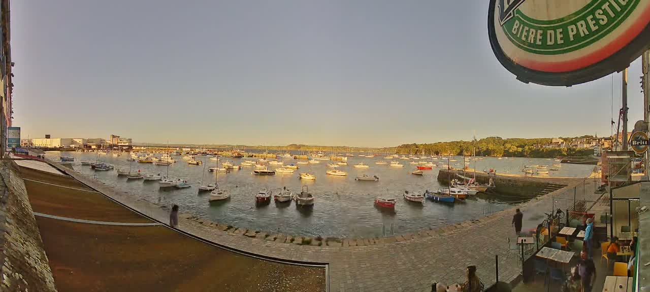 Numerous small boats are moored in a calm harbor at sunset, with a paved waterfront area and buildings visible in the background.