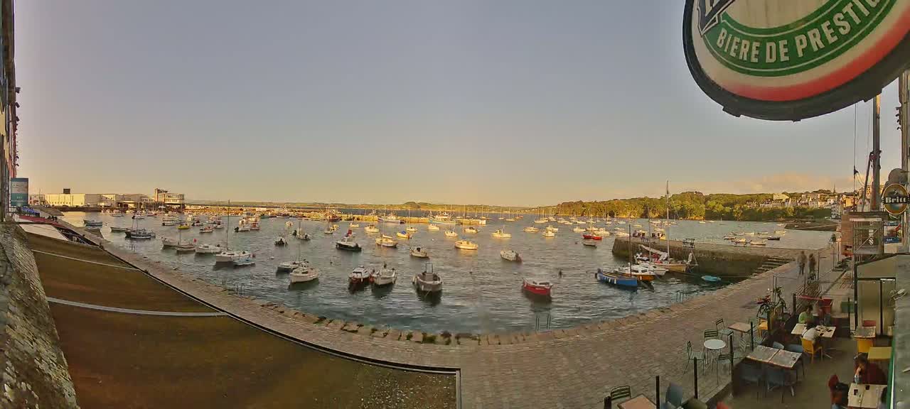 Many small boats are moored in a harbor at sunset, with a paved area and outdoor seating visible in the foreground.