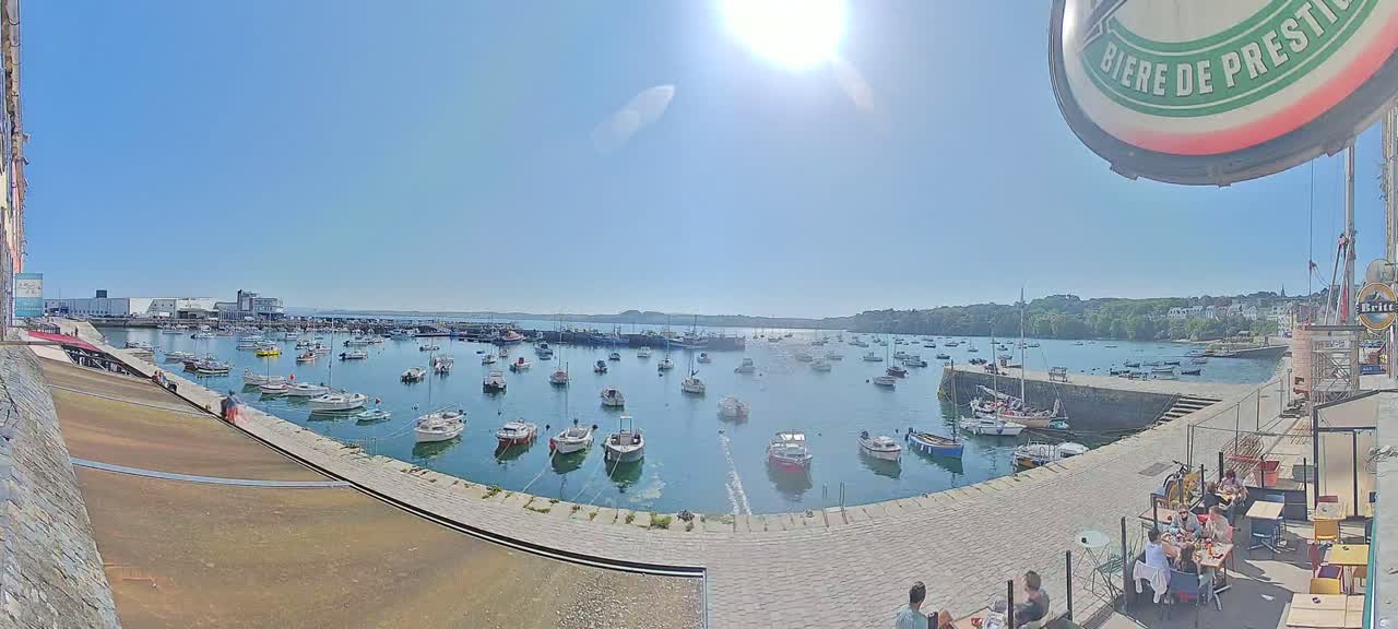 A sunny harbor filled with numerous boats, viewed from a paved area with outdoor seating.