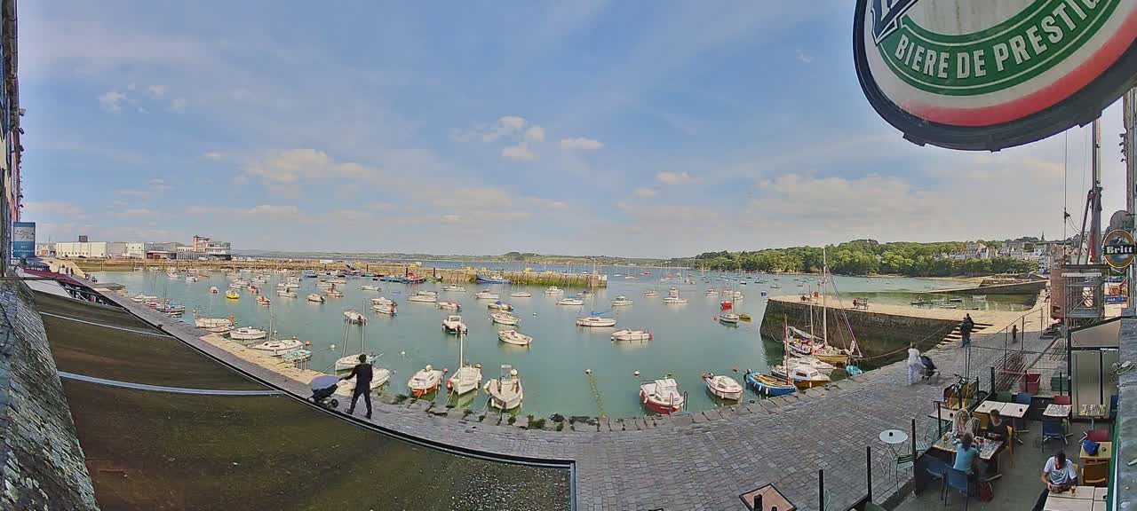 A harbor filled with numerous boats on a sunny day with partly cloudy skies is viewed from a cobblestone walkway next to a waterfront patio.
