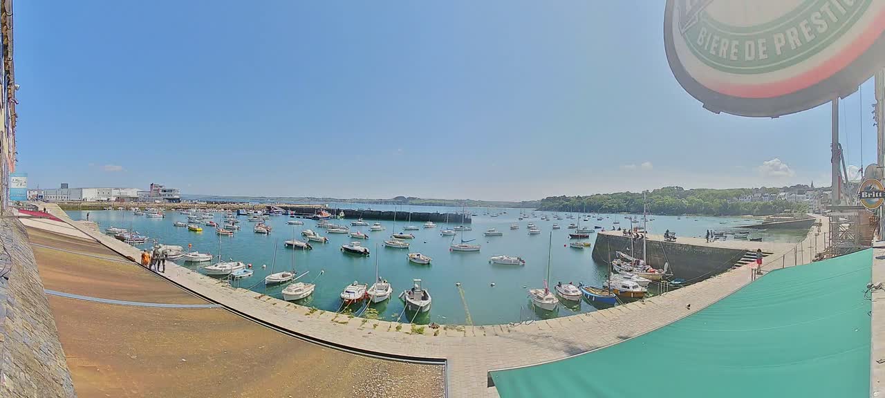 A sunny day shows a harbor filled with numerous small boats, nestled between stone walls and buildings, with green hills visible in the distance.