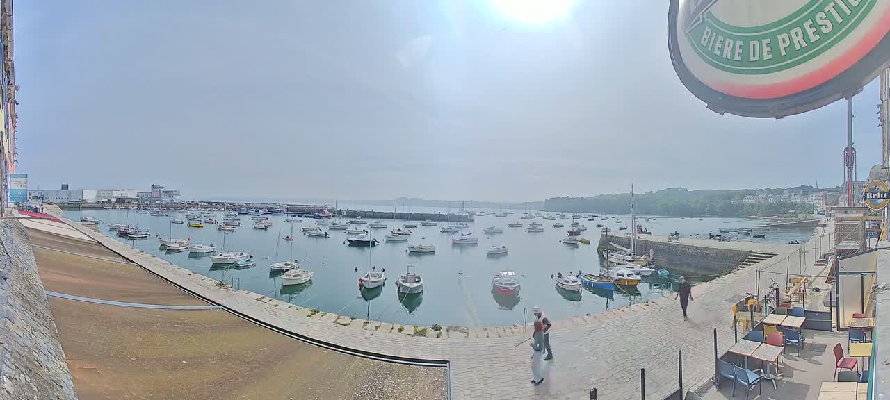 A sunny day shows a harbor filled with numerous small boats, a stone walkway with a few people, and outdoor seating at a nearby restaurant.