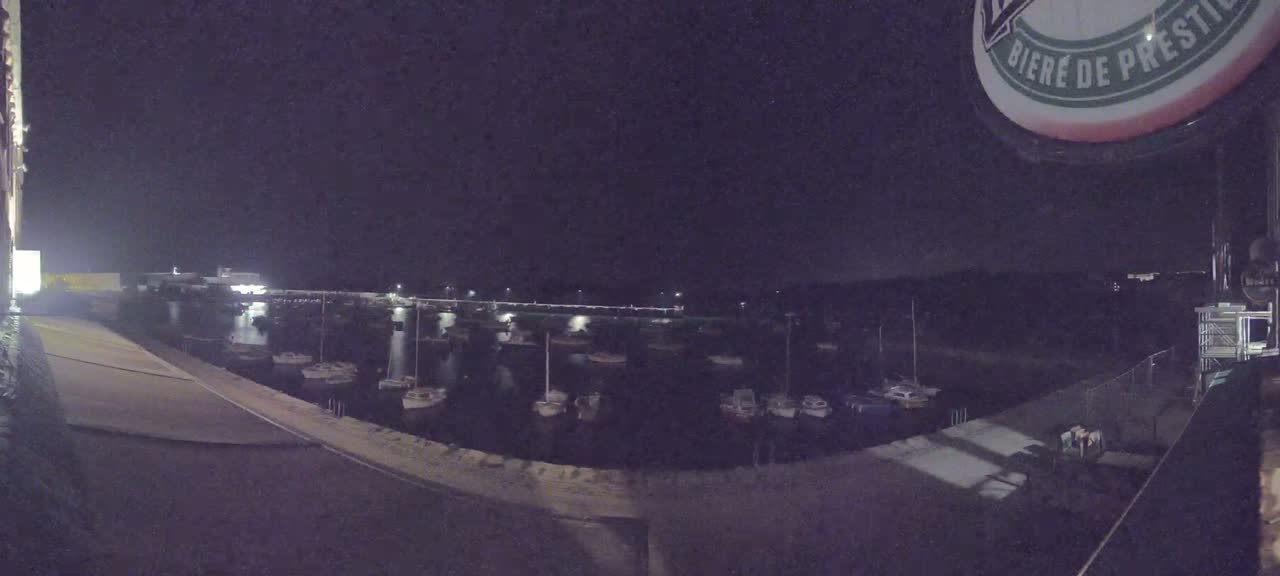 At night, numerous boats are moored in a calm harbor, illuminated by dim lights.