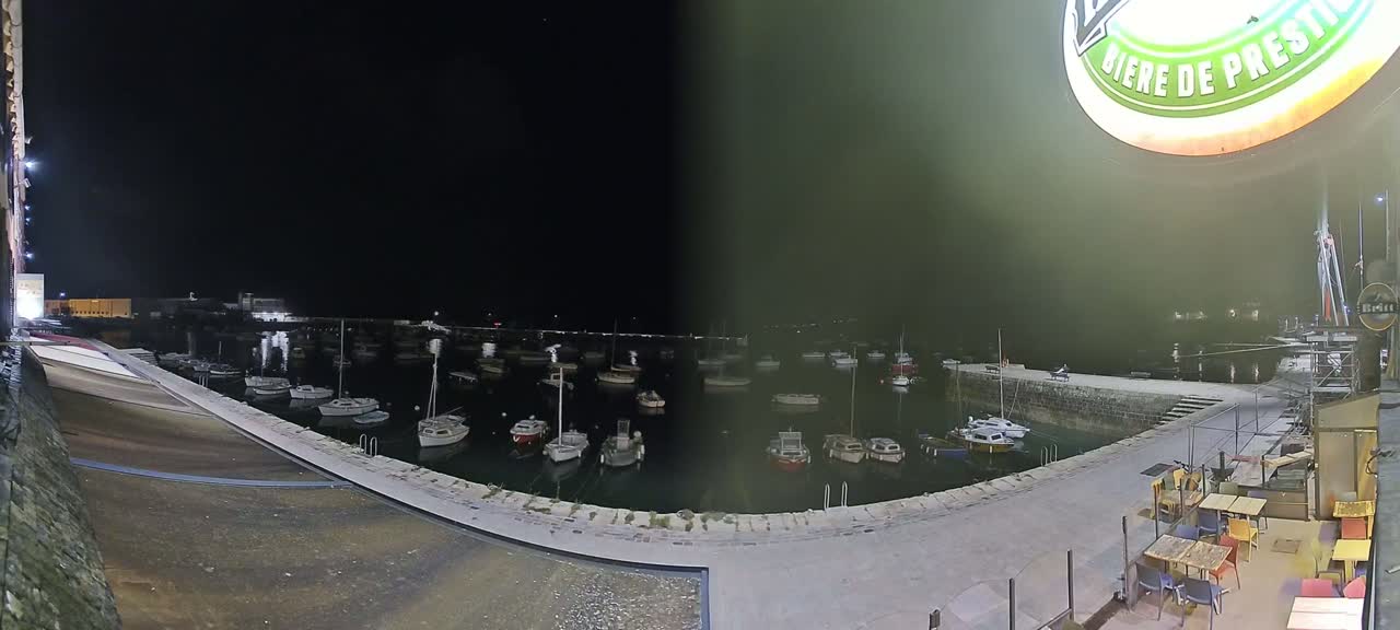 A nighttime view of a harbor filled with numerous small boats, seen from a slightly elevated vantage point overlooking a stone quay with outdoor seating.
