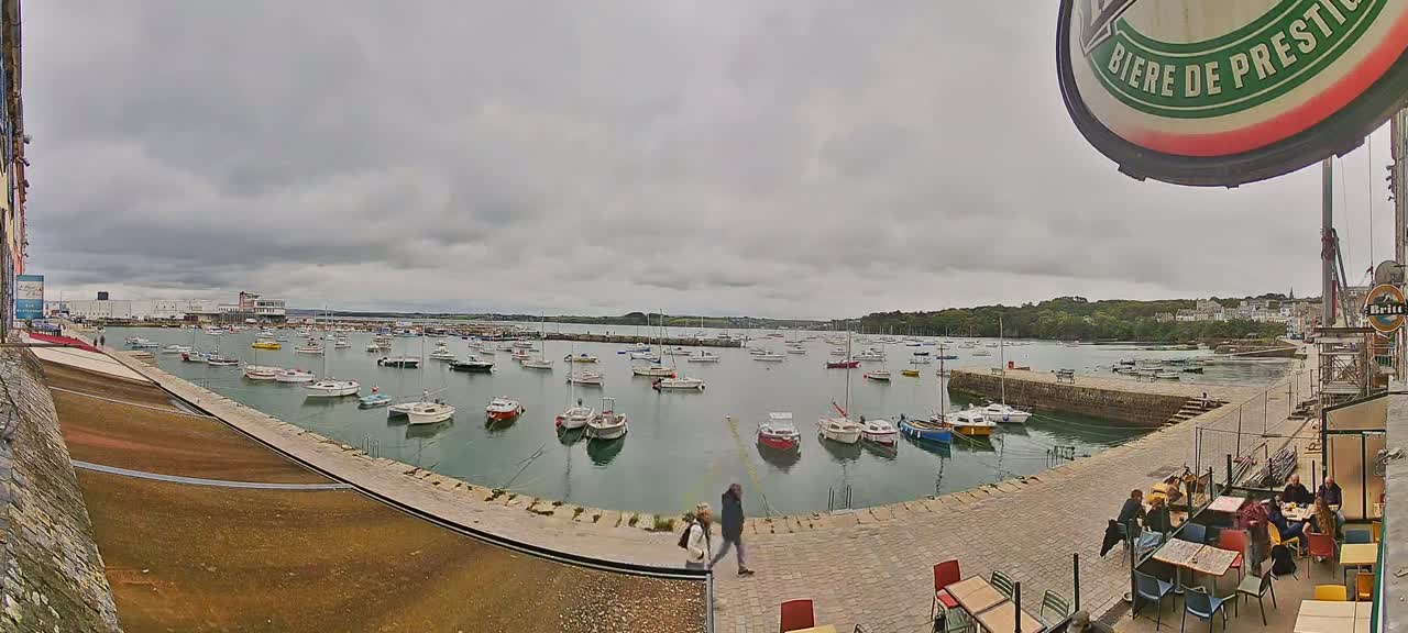 Many small sailboats are moored in a harbor on an overcast day, with people seated at outdoor tables in a nearby restaurant.