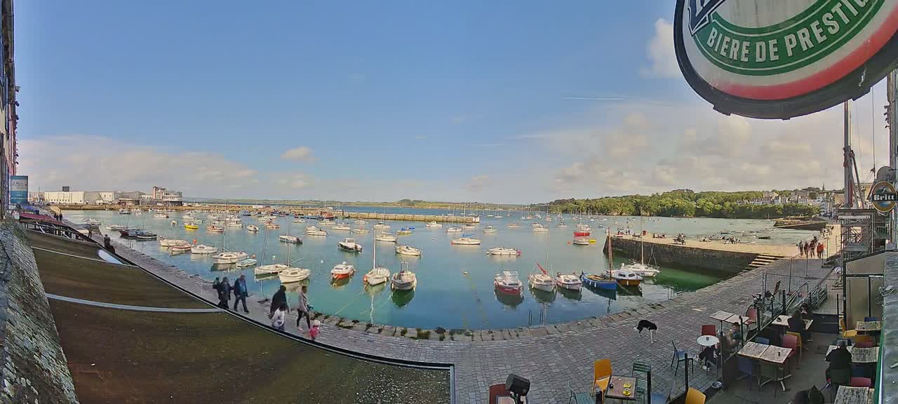 A harbor filled with numerous small boats under a partly cloudy, sunny sky, with people walking along a paved area near an outdoor cafe.