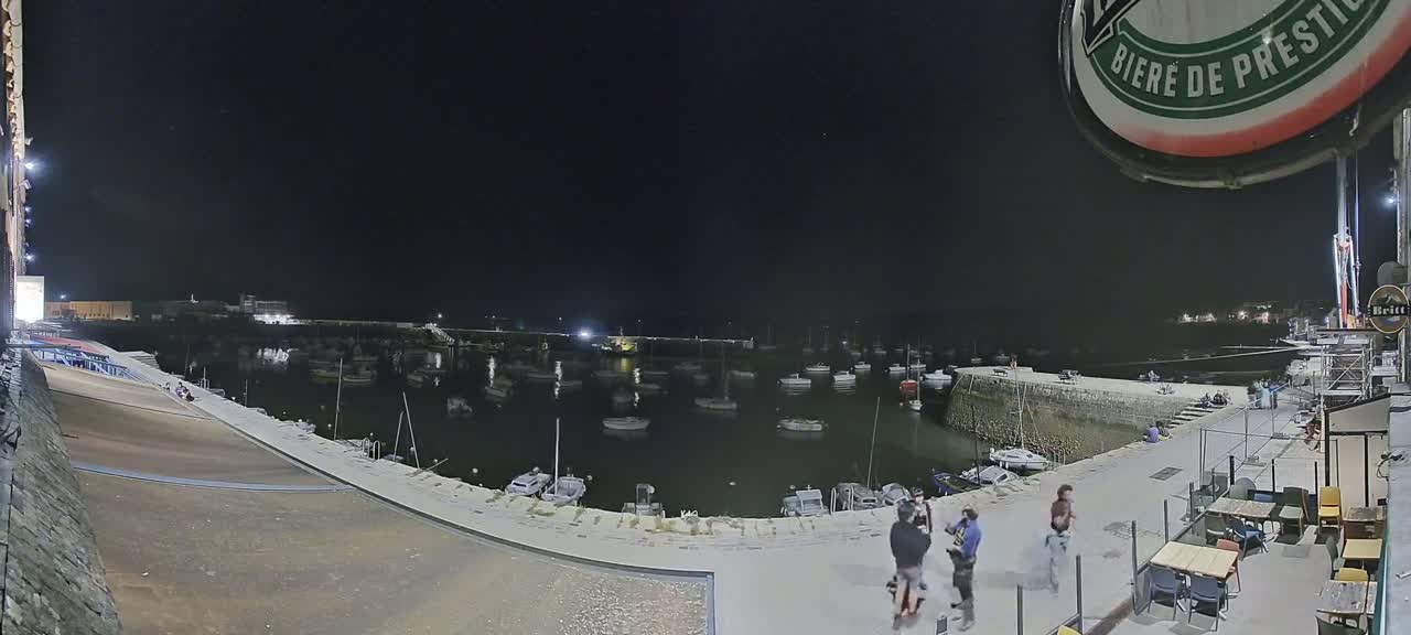 A nighttime harbor scene shows numerous boats moored in calm water, with a few people gathered on a nearby patio.