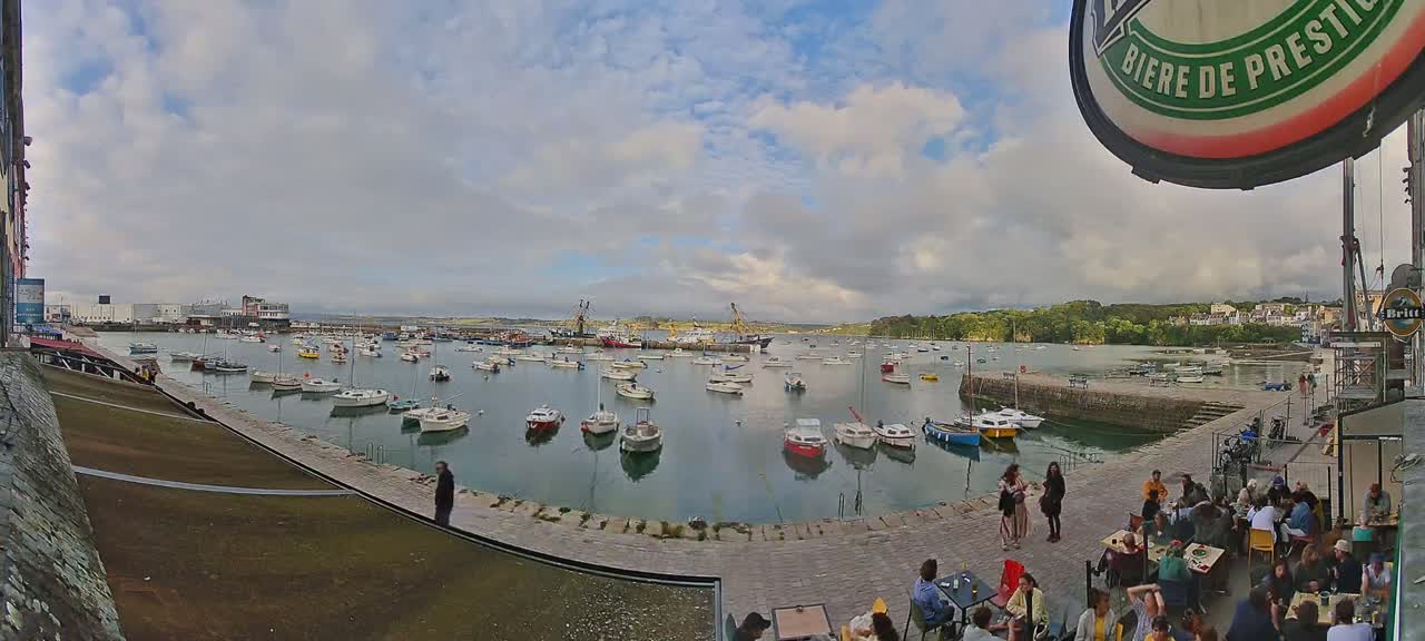 A harbor filled with numerous boats under a partly cloudy sky, with people sitting at outdoor tables along a stone walkway overlooking the water.
