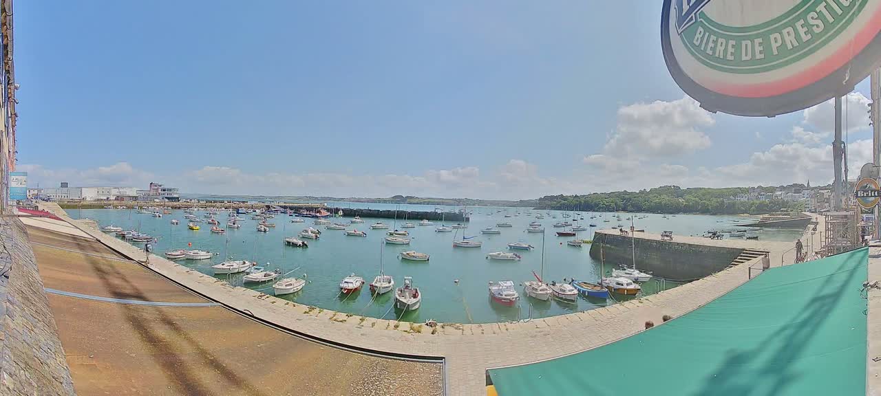 A sunny day shows numerous small boats moored in a harbor, with a stone quay and buildings visible in the background.