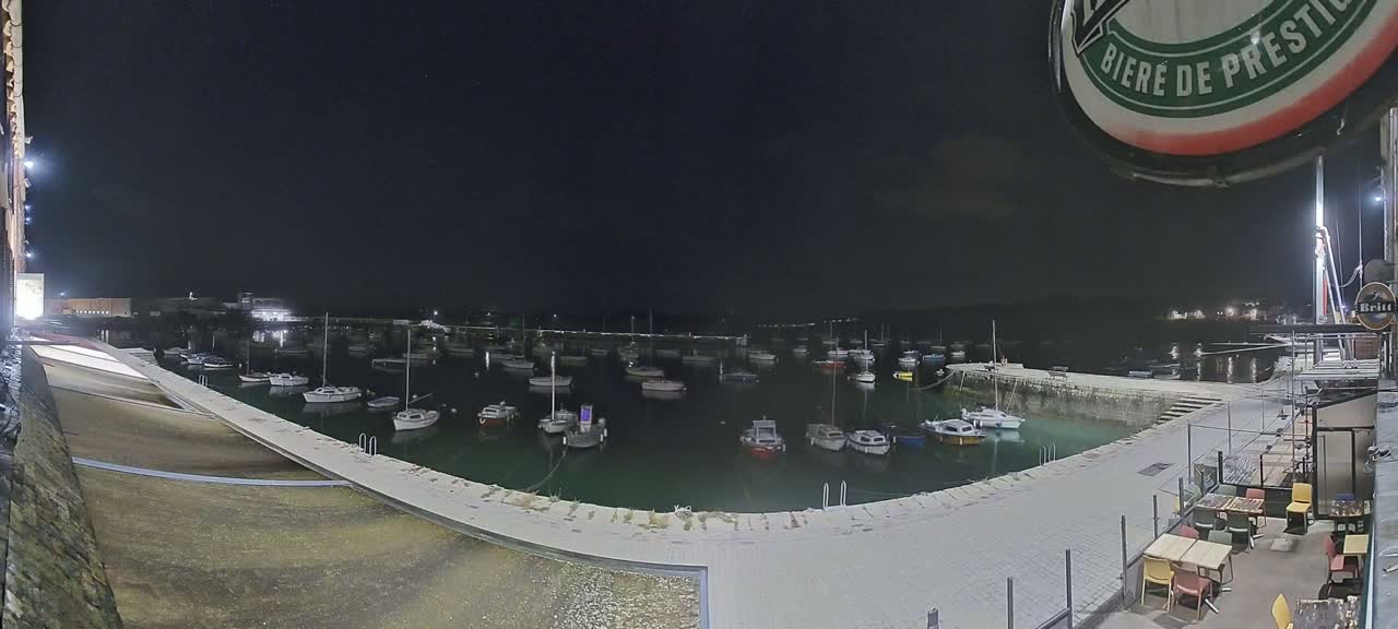 A nighttime view of a harbor filled with numerous boats moored along a stone quay, with outdoor seating at a nearby establishment under a clear, dark sky.