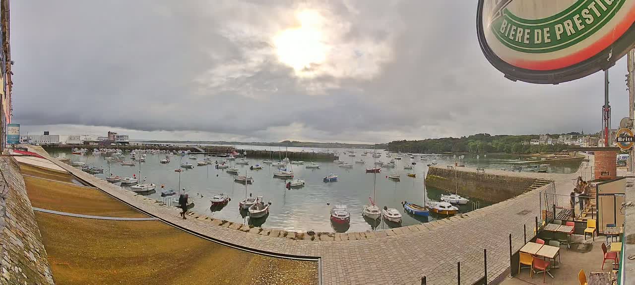 A harbor filled with numerous small boats is seen under a partly sunny, cloudy sky, with a paved walkway and outdoor seating area in the foreground.