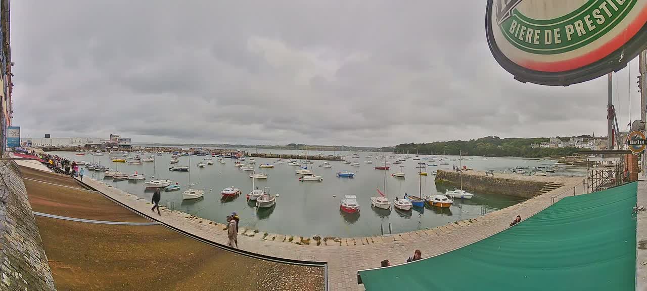 A harbor filled with numerous small boats is viewed from a quay on an overcast day, with people strolling along the waterfront.